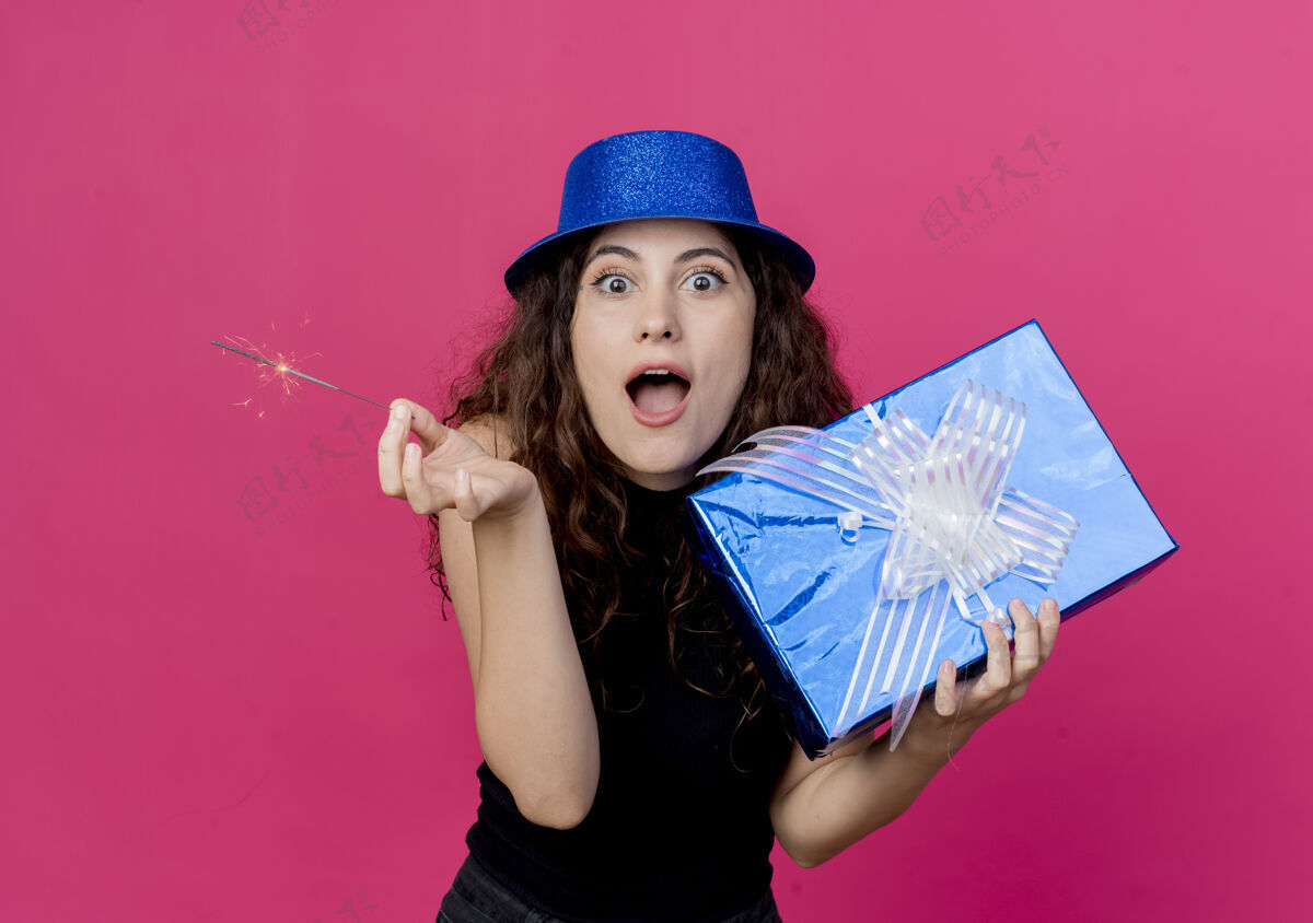站年轻漂亮的女士 卷发 戴着节日礼帽 手里拿着生日礼盒和闪闪发光的惊喜 生日派对的概念超过了粉色帽子闪闪发光生日