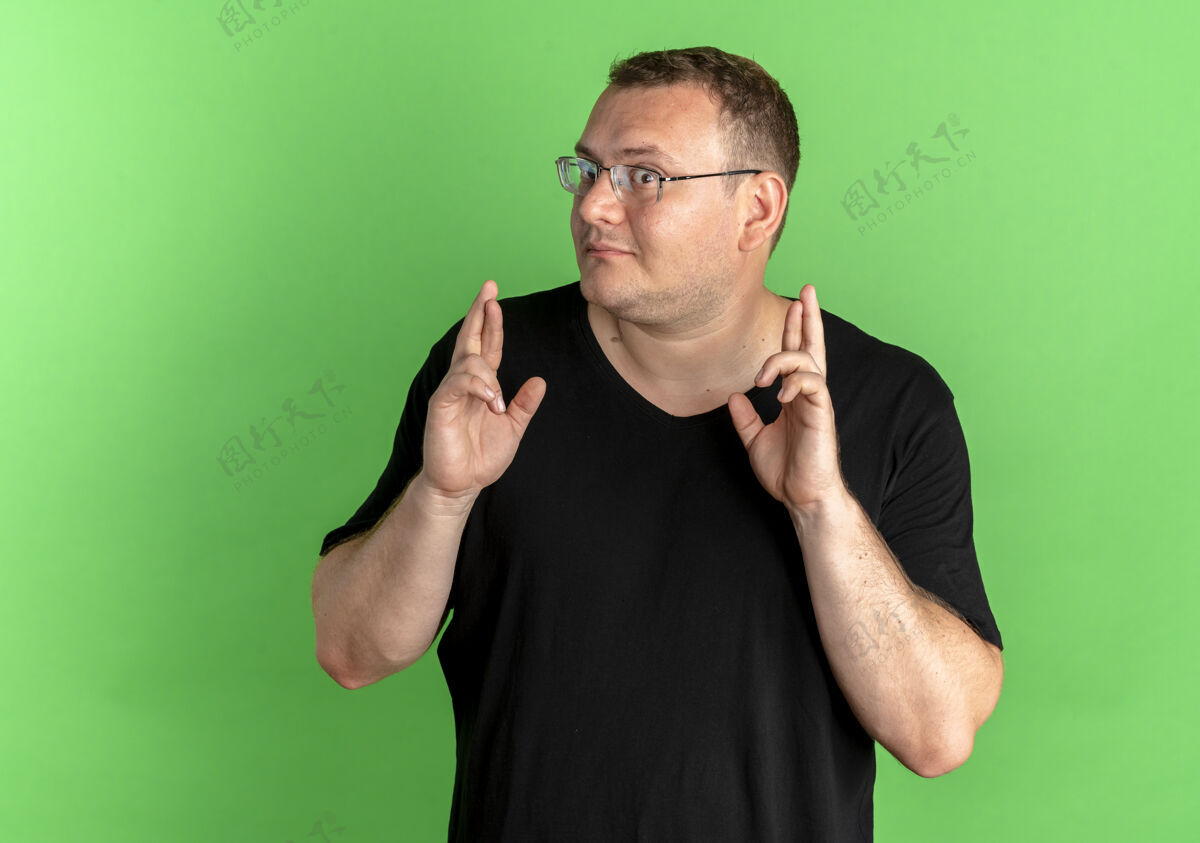 脸戴眼镜的超重男子穿着黑色t恤许下美好的愿望手指交叉 脸上露出幸福的绿色愿望超重穿