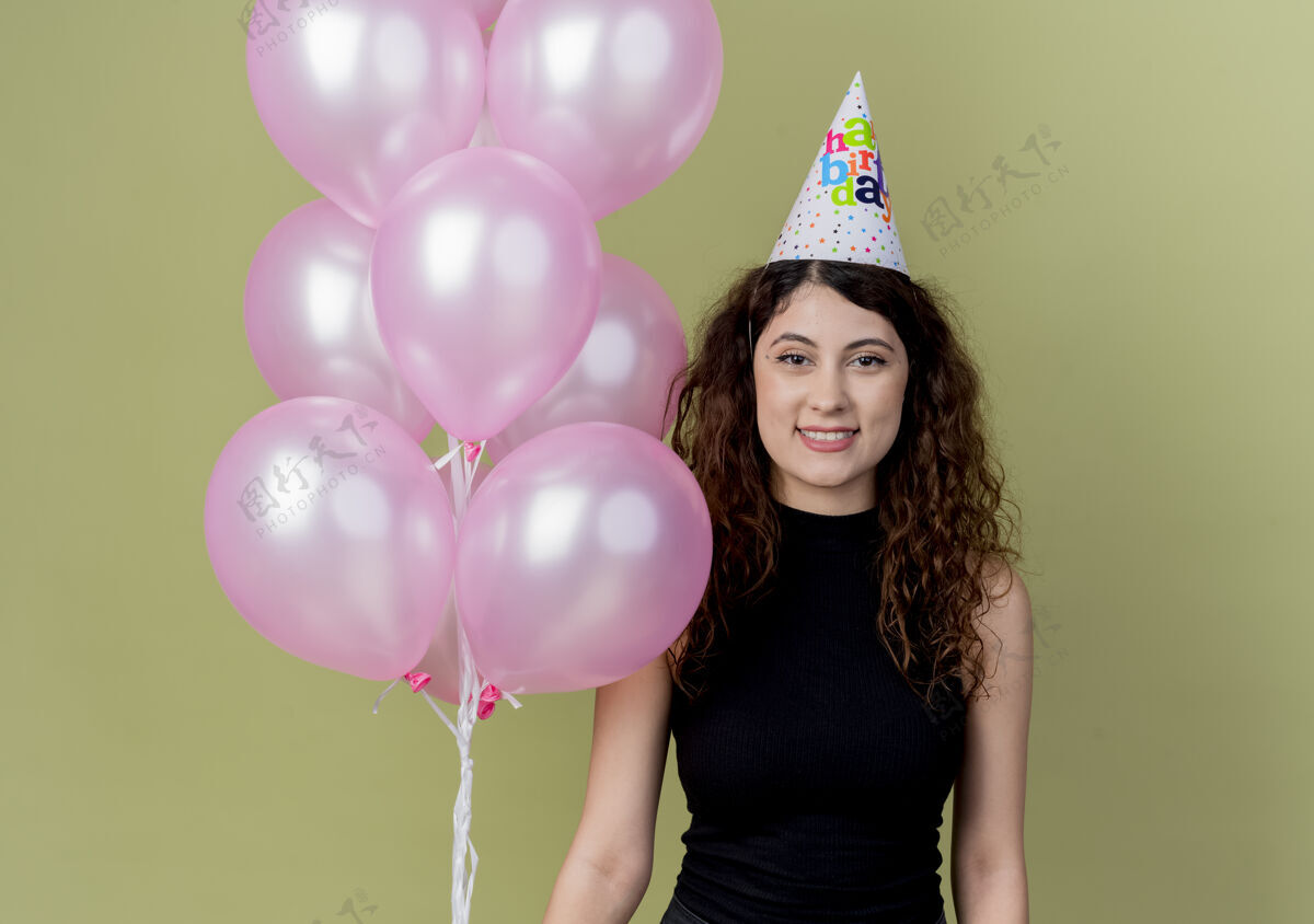 年轻戴着节日帽 留着卷发的年轻漂亮女人手持气球看着镜头 在灯光下微笑着 快乐而积极头发看卷曲