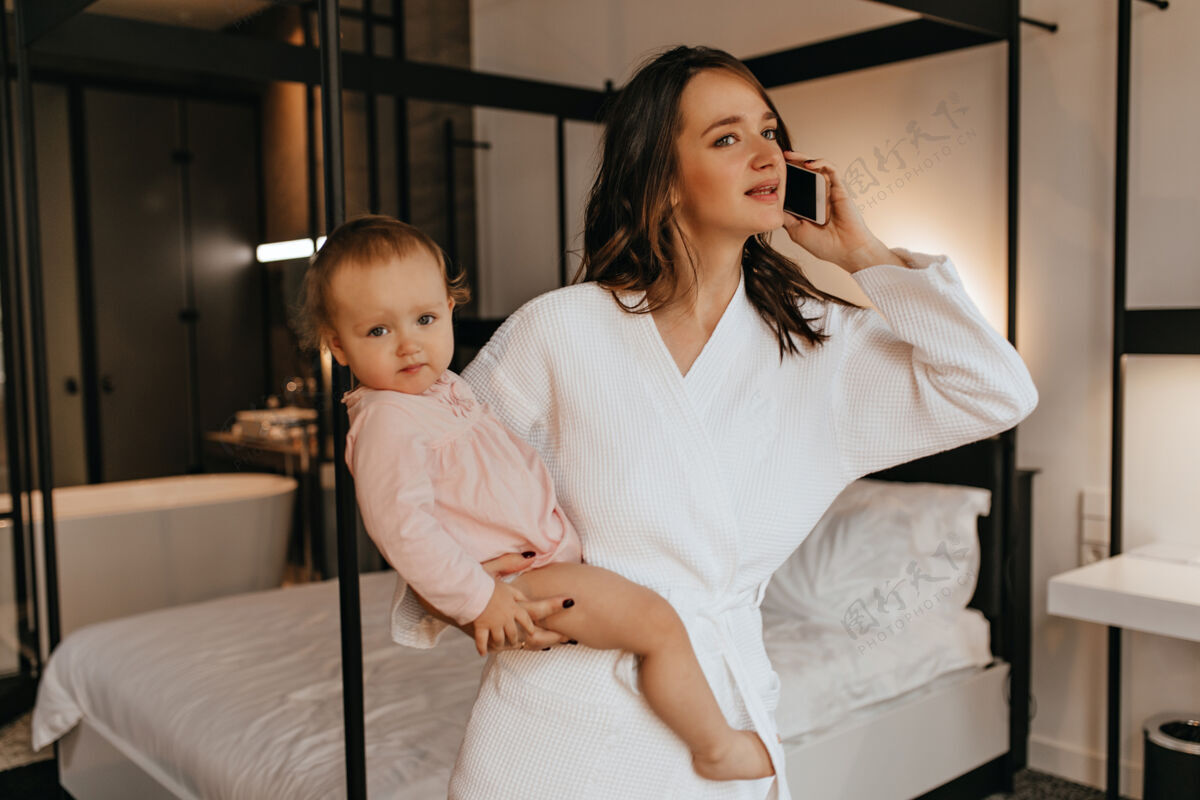 房间年轻漂亮的母亲穿着白色浴袍 抱着女儿 在卧室里打电话爱女性父母
