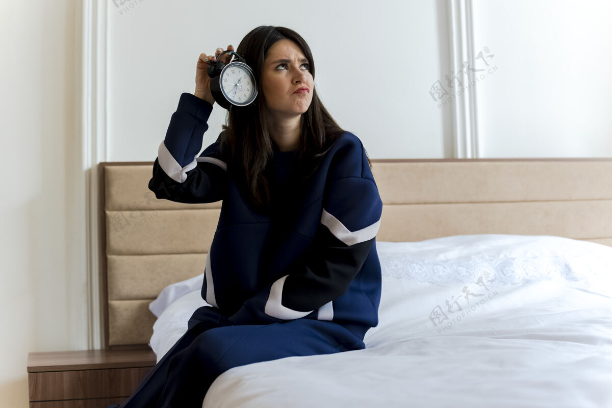 女人一个年轻漂亮的白人女人坐在卧室的床上 拿着闹钟在床头边看人侧身黑发