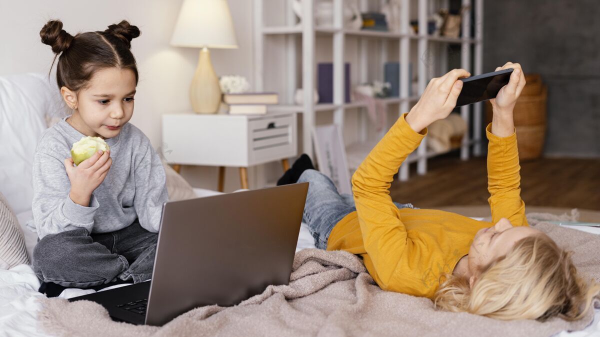 室内兄弟姐妹在床上看笔记本电脑和手机上的视频房子女孩苹果