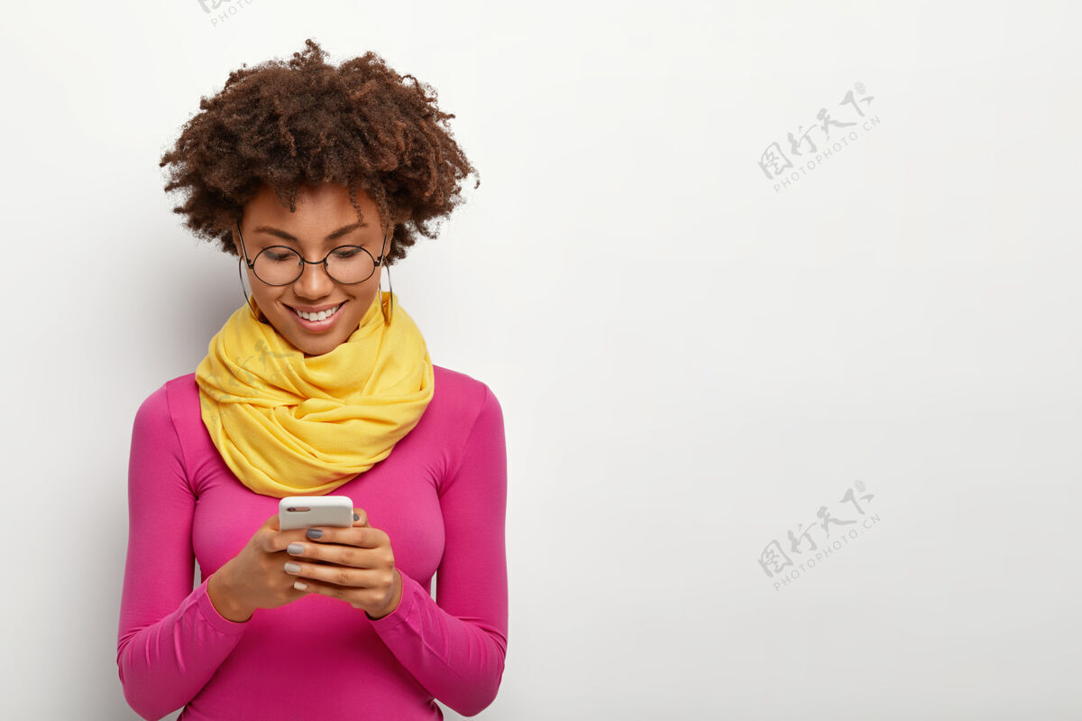 深色头发喜得皮肤黝黑的女性手持现代手机 专注于展示 穿着粉色高领毛衣眼镜人非洲