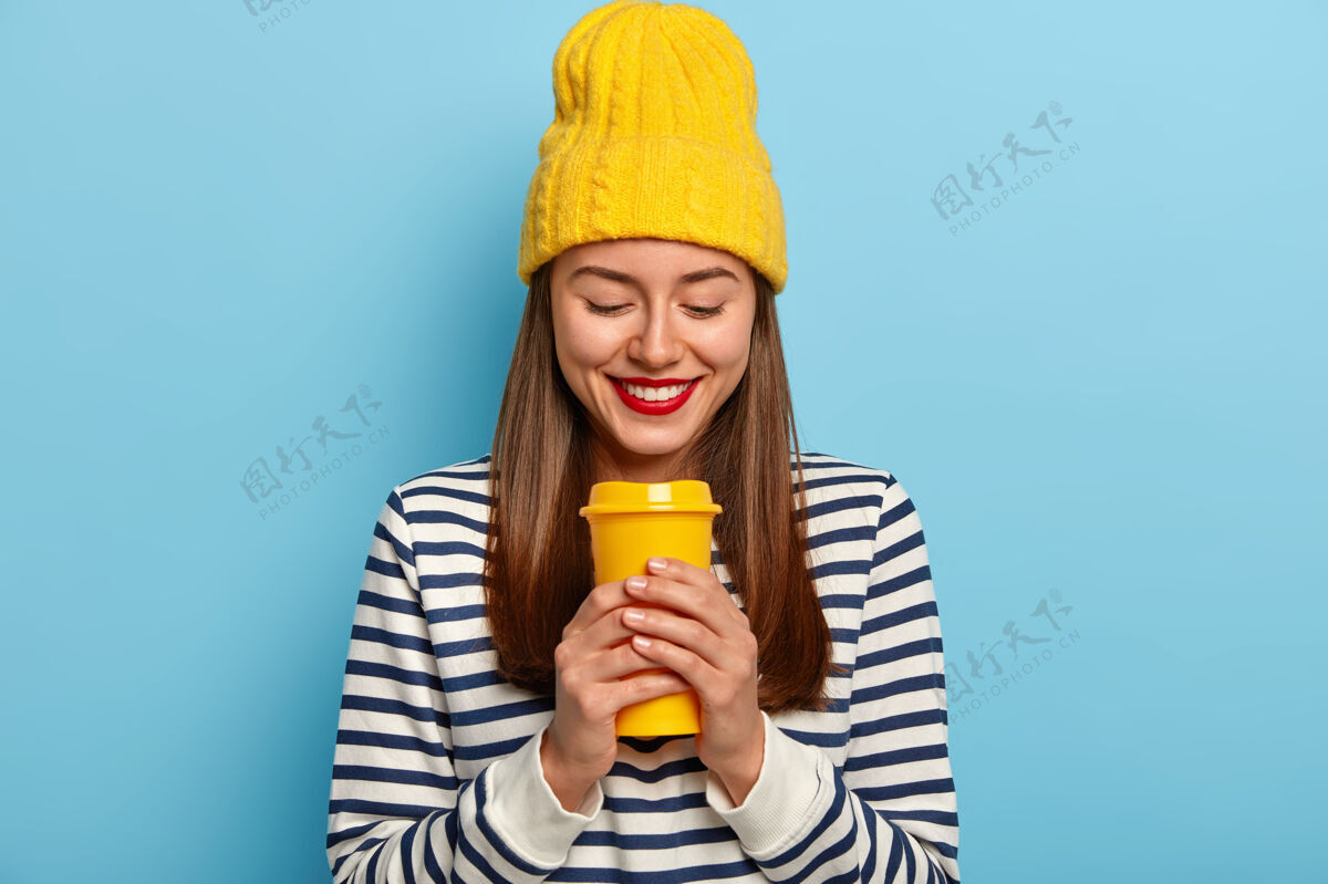 化妆品快乐的女人戴着时髦的黄色帽子和条纹套头衫 拿着外卖咖啡 涂着红色的嘴唇 享受着芳香的饮料自由时间站立外卖