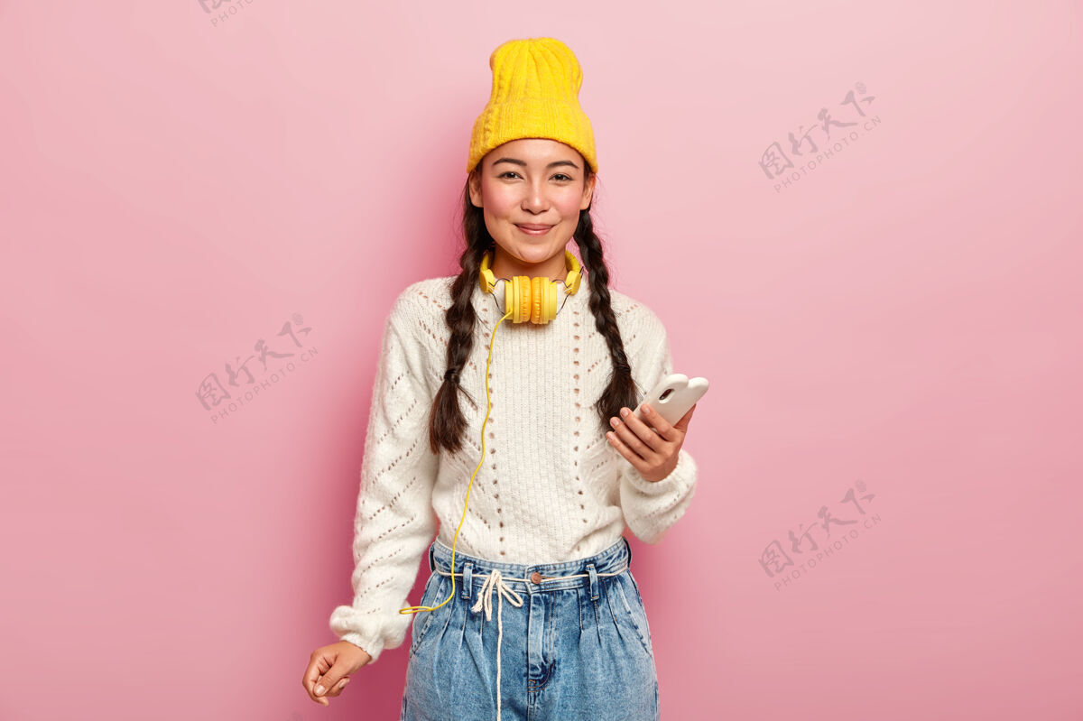 休息可爱的黑头发小辫子女孩 用手机上网 戴着时髦的头饰帽子设备毛衣