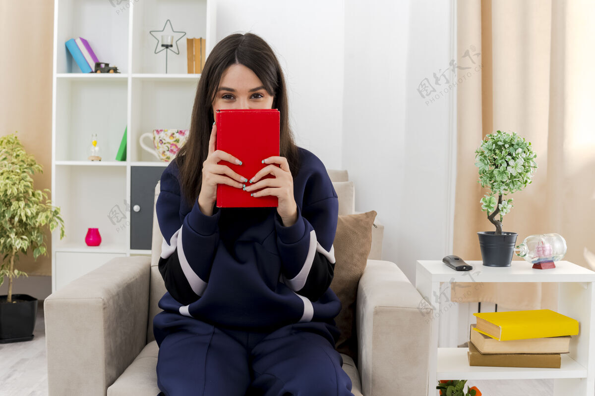 看年轻漂亮的白人妇女坐在设计客厅的扶手椅上 手里拿着书 从书的后面看抱着住着书