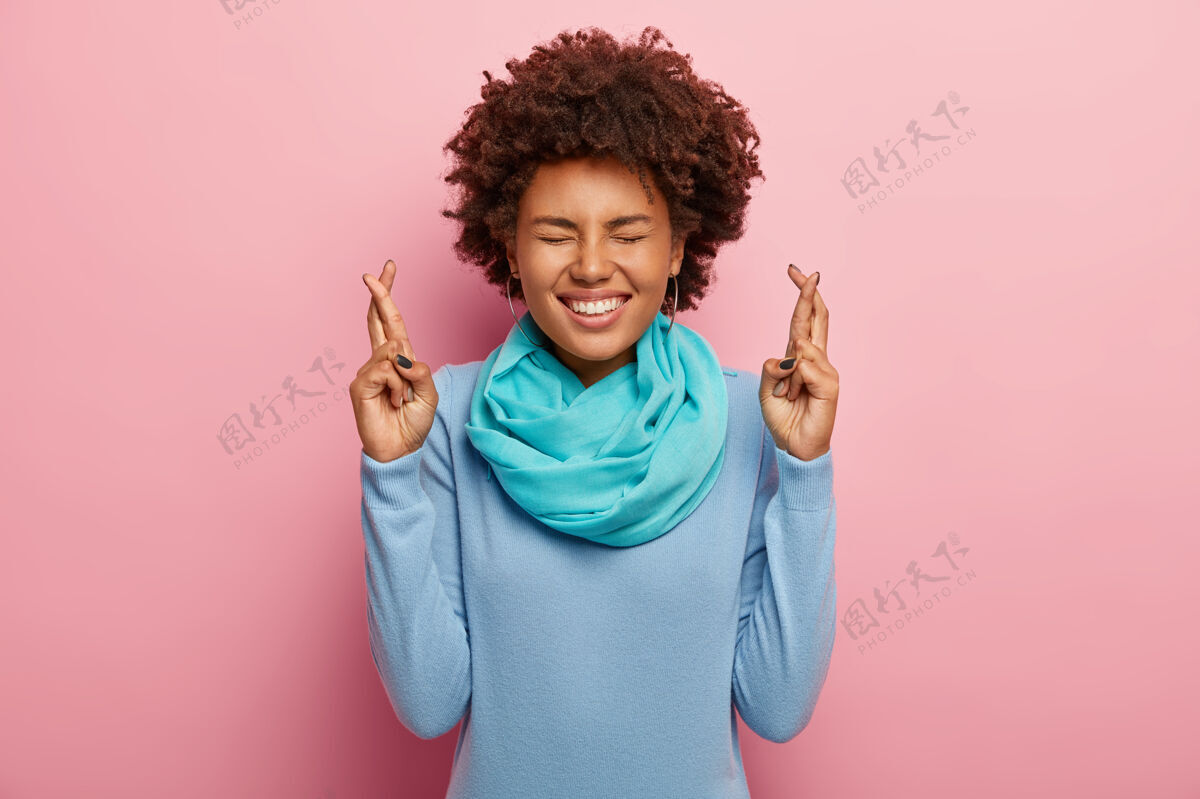 年轻人喜出望外的非洲头发女人的肖像 手指交叉 相信好运 笑容灿烂 穿着蓝色毛衣和围巾快乐人欣喜若狂