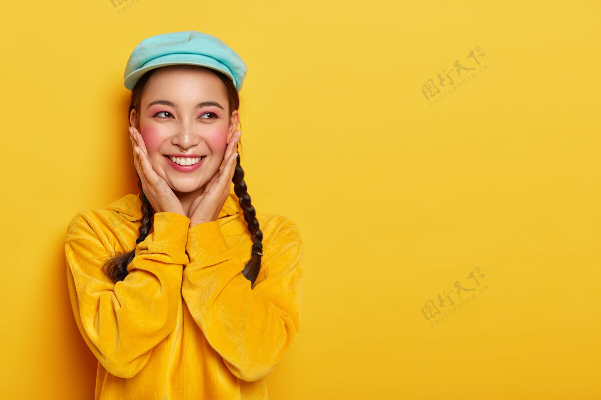 快乐快乐的亚洲女孩抚摸着胭脂的脸颊 化着妆 穿出刺眼 时髦的帽子和丝绒运动衫 有着梦幻般的表情亚洲人肤色模特