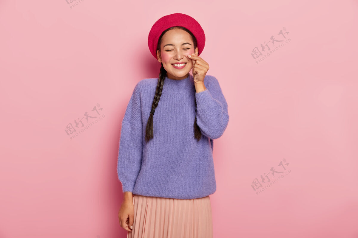 手臂快乐快乐的亚洲女人用手塑造小心形 做韩式招牌 头戴红色贝雷帽 穿着休闲套头衫和裙子 微笑愉快 心情愉快 隔着粉色的墙与世隔绝快乐女性编织