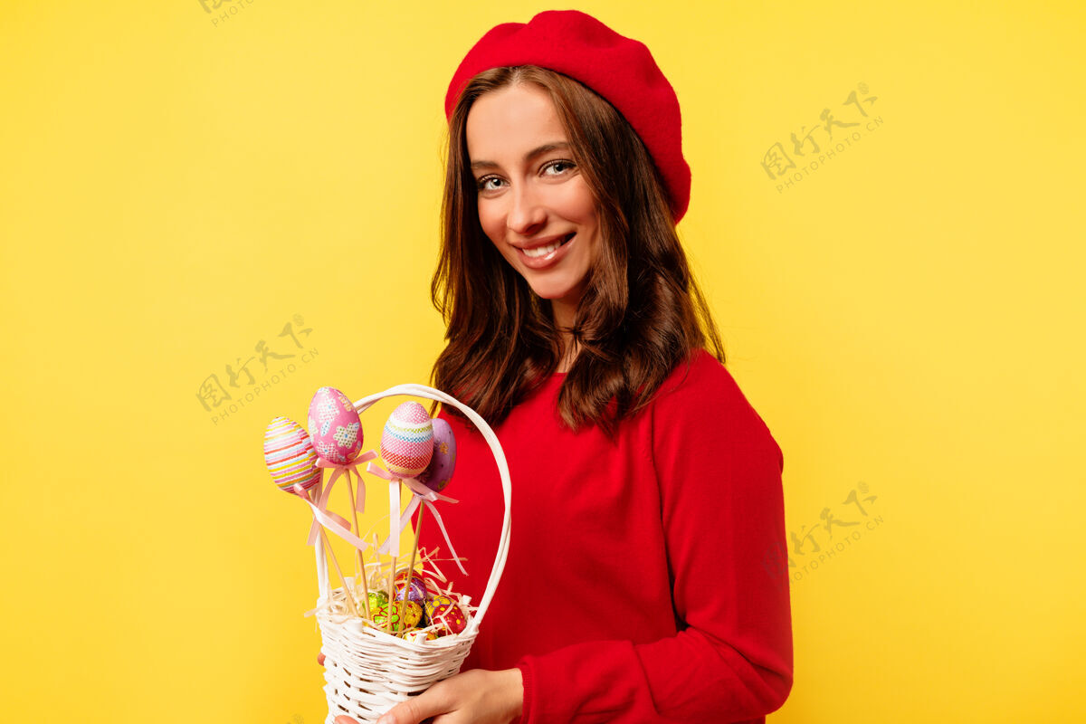 魅力快乐微笑的漂亮女人 卷发 穿着红色套头衫 头戴红色贝雷帽 在隔离的黄色墙壁上摆着复活节篮子的姿势笑贝雷帽微笑