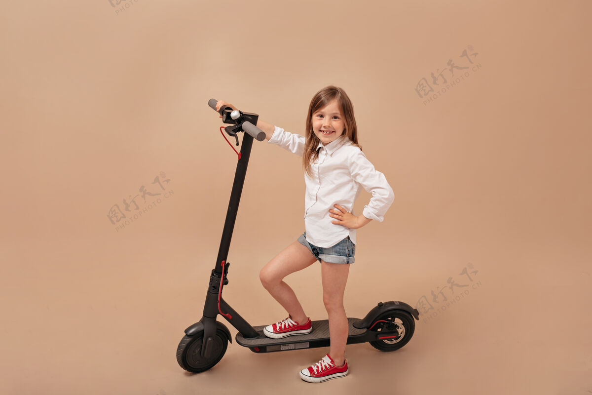 享受室内框架的小可爱的女孩在孤立的米色背景与电动滑板车相机摆姿势幼儿年轻易趣