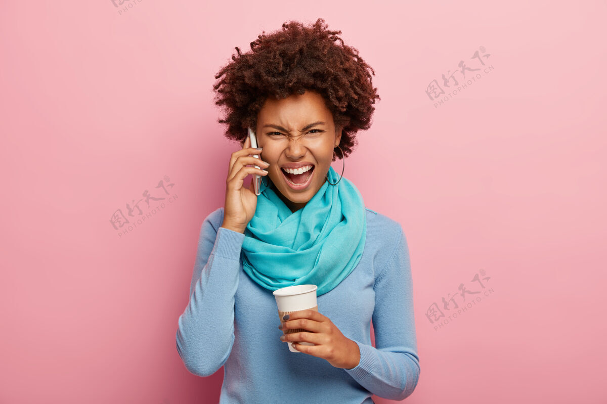 喊叫恼怒的非洲女人通过智能手机吵架 沮丧地大声喊叫 拿着外卖咖啡杯 戴着蓝色套头衫和围巾电话抱着人