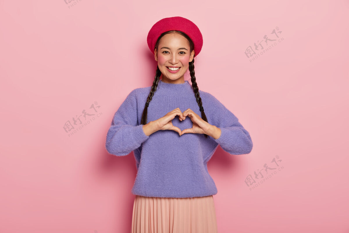 心外表迷人的年轻女士 在胸前做一个心形手势 承认爱情 戴着红色贝雷帽 紫色超大套头衫 化着妆 隔离在粉红色的墙上微笑套头衫休闲
