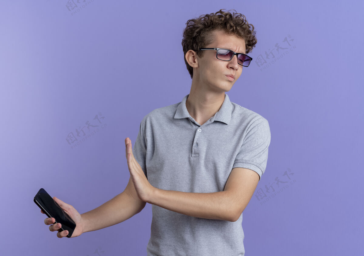 眼镜一个戴着黑眼镜 穿着灰色马球衫 手持智能手机的年轻人站在蓝色的墙上做着防御的手势手年轻的握着