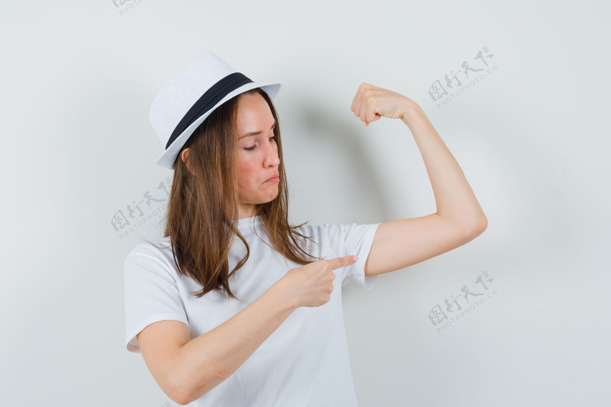 年轻穿着白色t恤的年轻女孩 帽子指着手臂的肌肉 看上去很自豪 前视图童年小指向