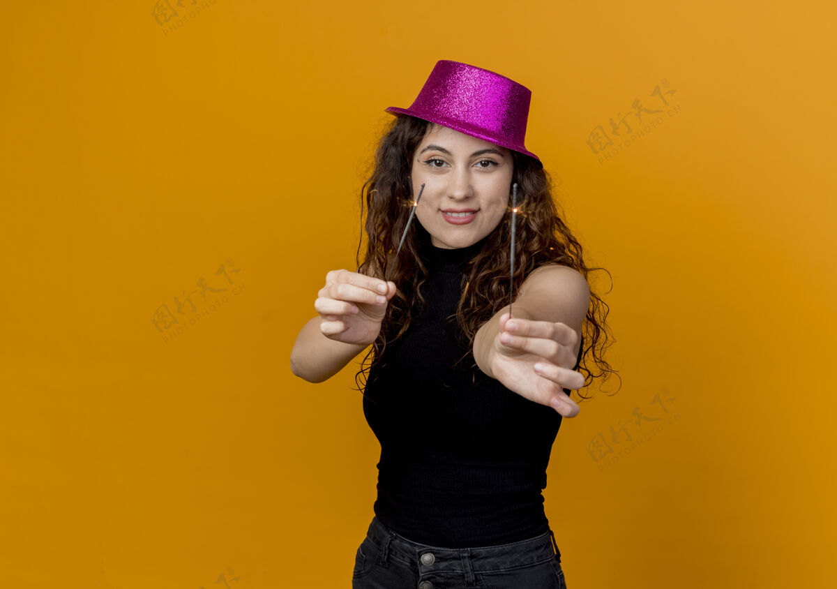女人一个年轻漂亮的女人 卷发 戴着派对帽 手里拿着火花 站在橘色的墙上 快乐而兴奋卷发帽子年轻