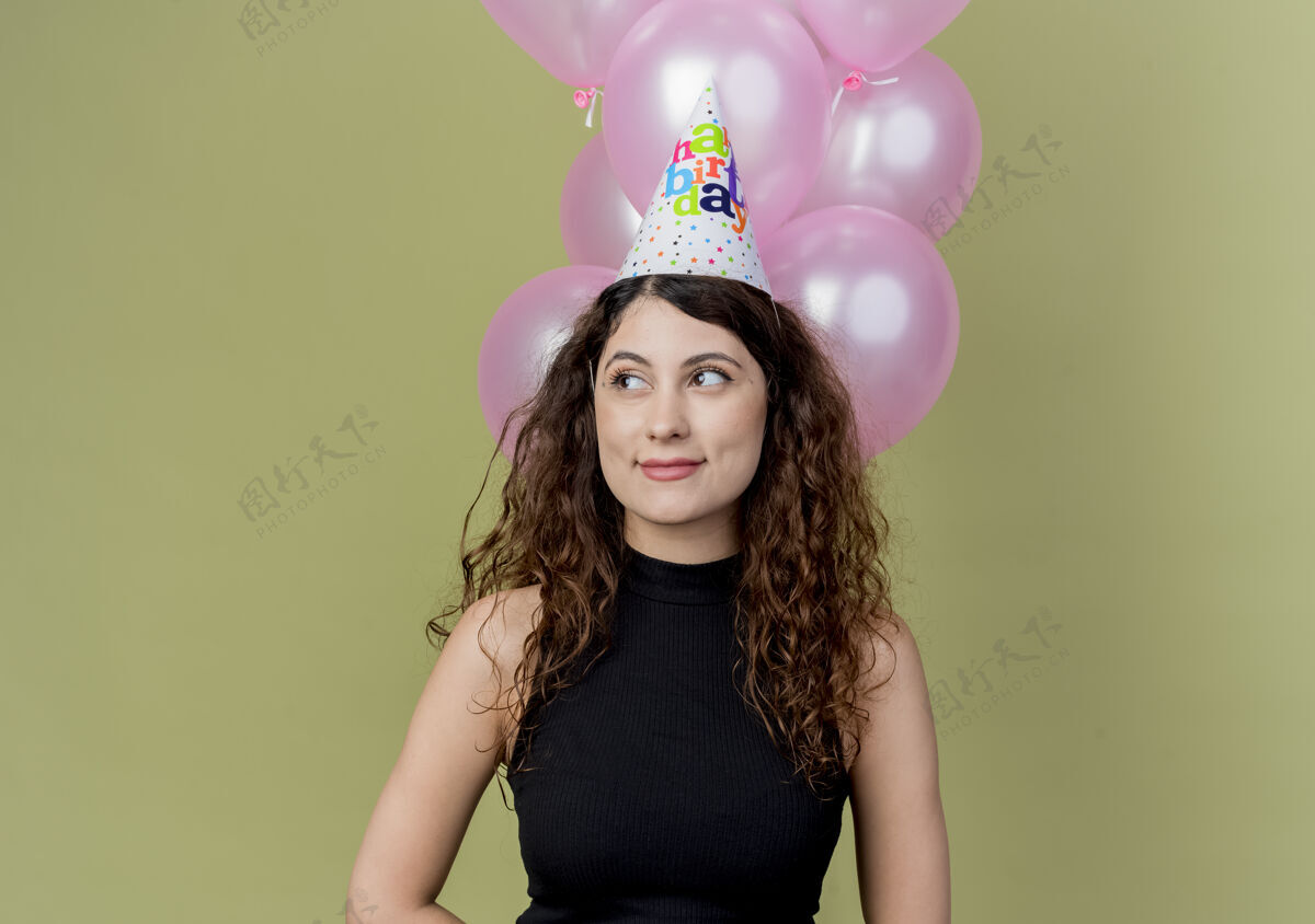微笑一个年轻漂亮的女人 卷发 戴着节日帽 手里拿着气球 脸上带着微笑 站在光墙上 生日派对的概念光看女人