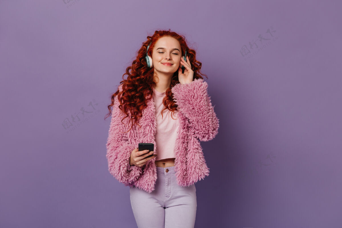 女性穿着时尚的粉色和白色套装 戴着蓝色耳机欣赏音乐的平静女孩红头发的女人在淡紫色的空间里和智能手机合影蓝色眼睛女性化粉色外套