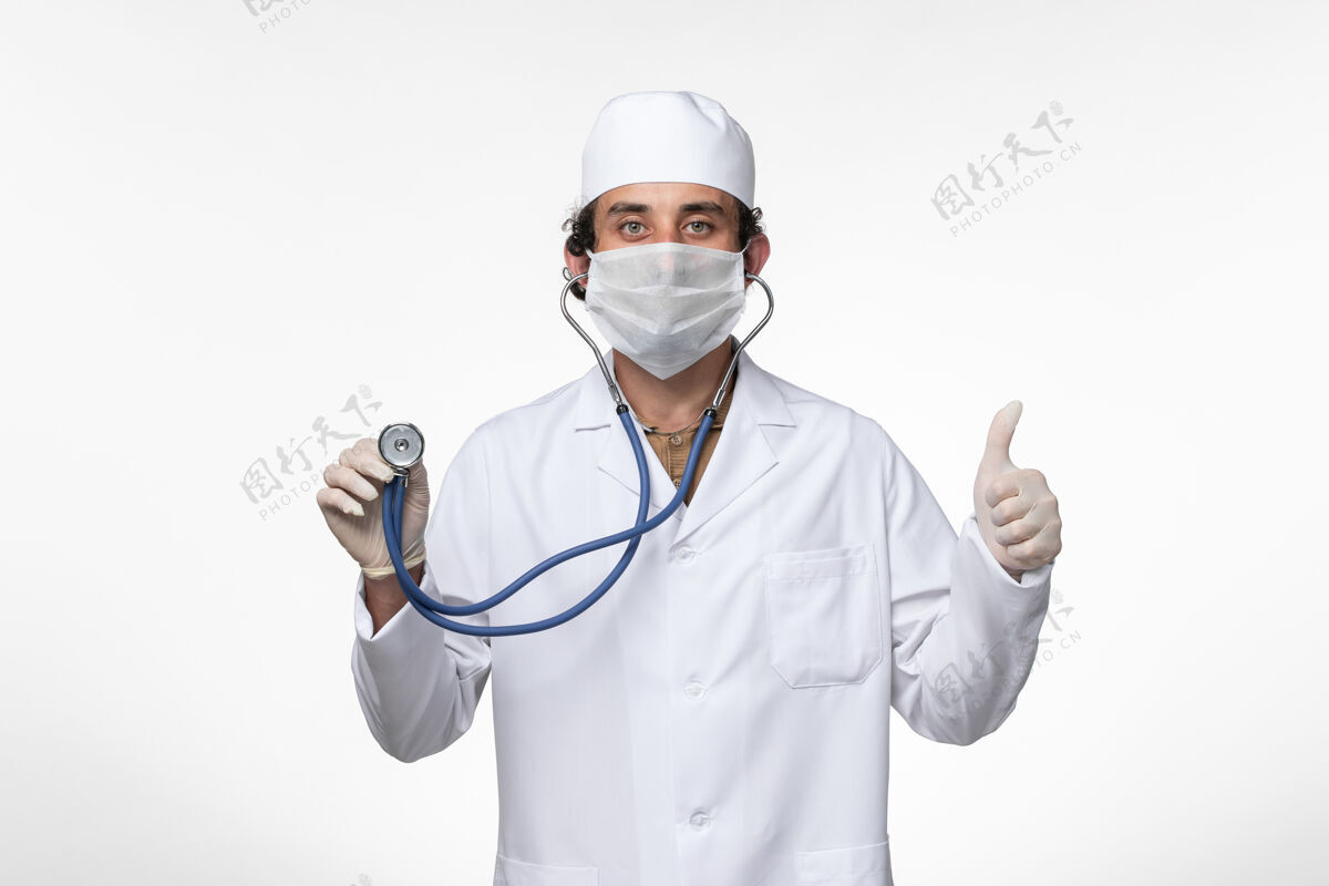 使用正面图：男医生穿着医疗服 戴着口罩以防病毒感染-使用听诊器对白色桌面疾病病毒进行检测-病毒感染-大流行医疗外套冠状病毒预防