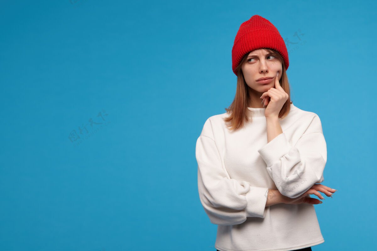 情绪严肃的 深色头发的成年女孩的肖像穿着白色毛衣和红色帽子用手指触摸她的脸颊和思考在复制空间的左边看 隔离在蓝色的墙上外观脸颊思考