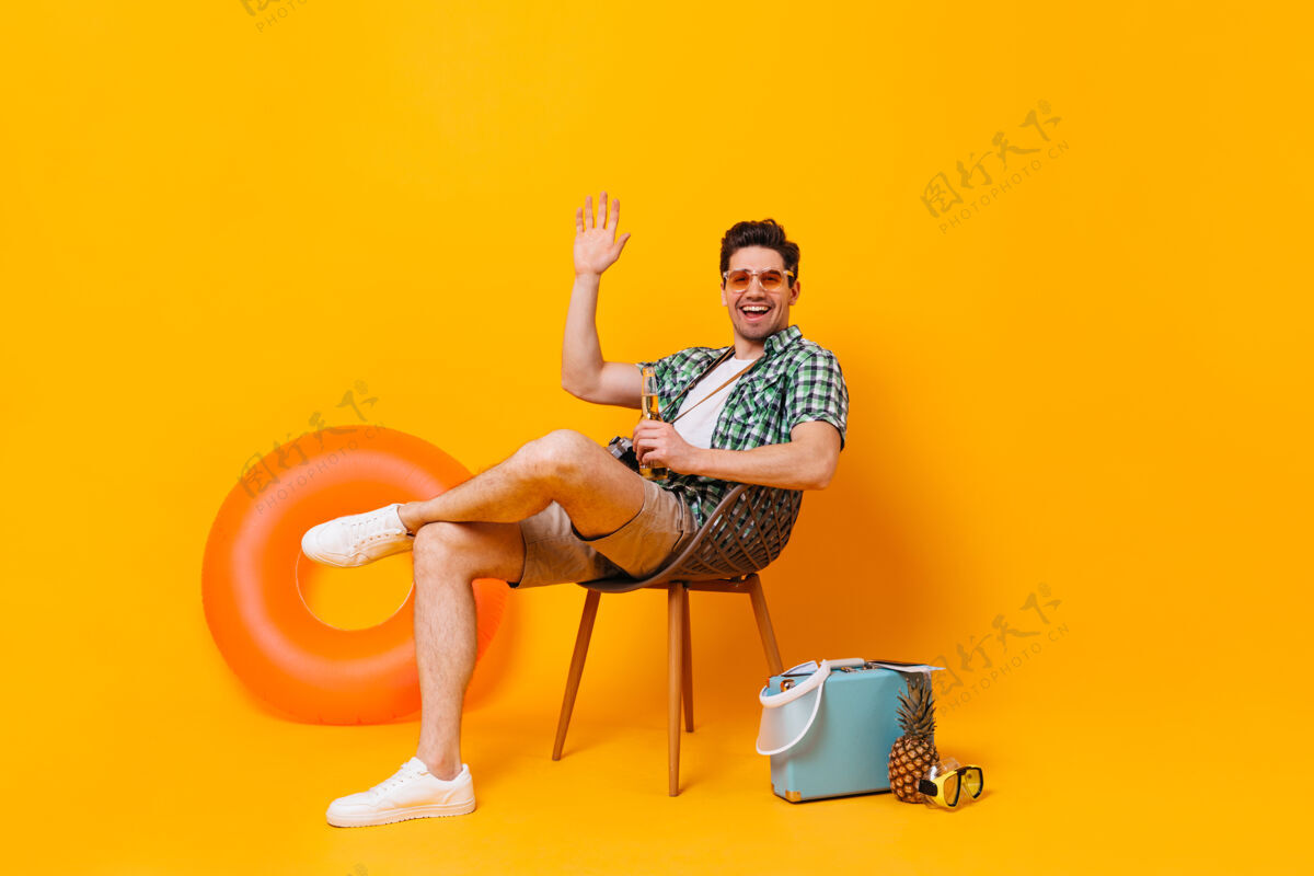 行李戴着墨镜的年轻人在充气圈和手提箱的衬托下享受假期那个家伙正坐在木椅上 喝着啤酒 挥舞着手信心冒险椅子