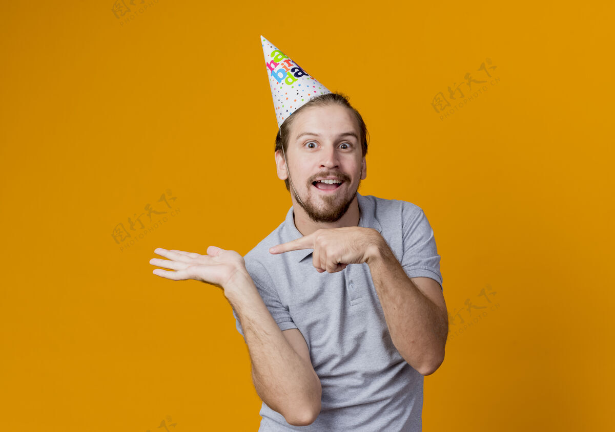 侧身戴节日帽的年轻人伸出手臂 用手指着站在橘色墙上的生日派对概念手臂男人站