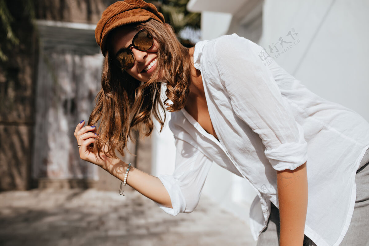 太阳镜胳膊上有漂亮纹身的卷发女人在笑穿着白衬衫和白帽子的友好女孩在舒适的房子里看着摄像机女士华丽放松