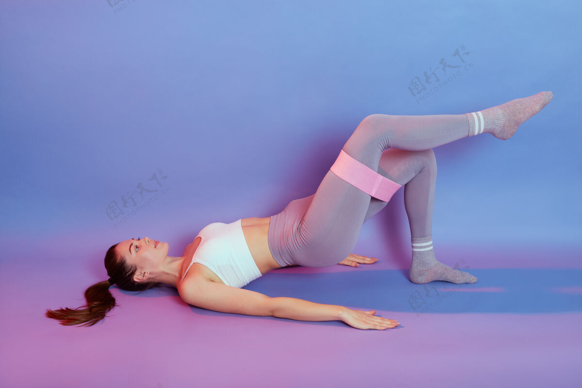 肌肉迷人的运动型女士 完美的身体摆在地板上 做臀部练习 抬起一只手 使用松紧带 在彩色背景下独立锻炼身体强壮手臂