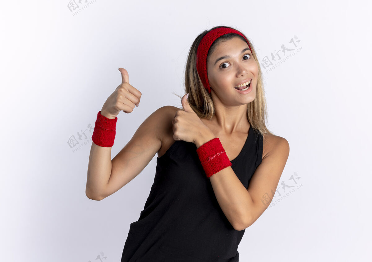 秀身穿黑色运动服 头戴红色头巾的年轻健身女孩站在白墙上微笑着竖起大拇指微笑运动装站立