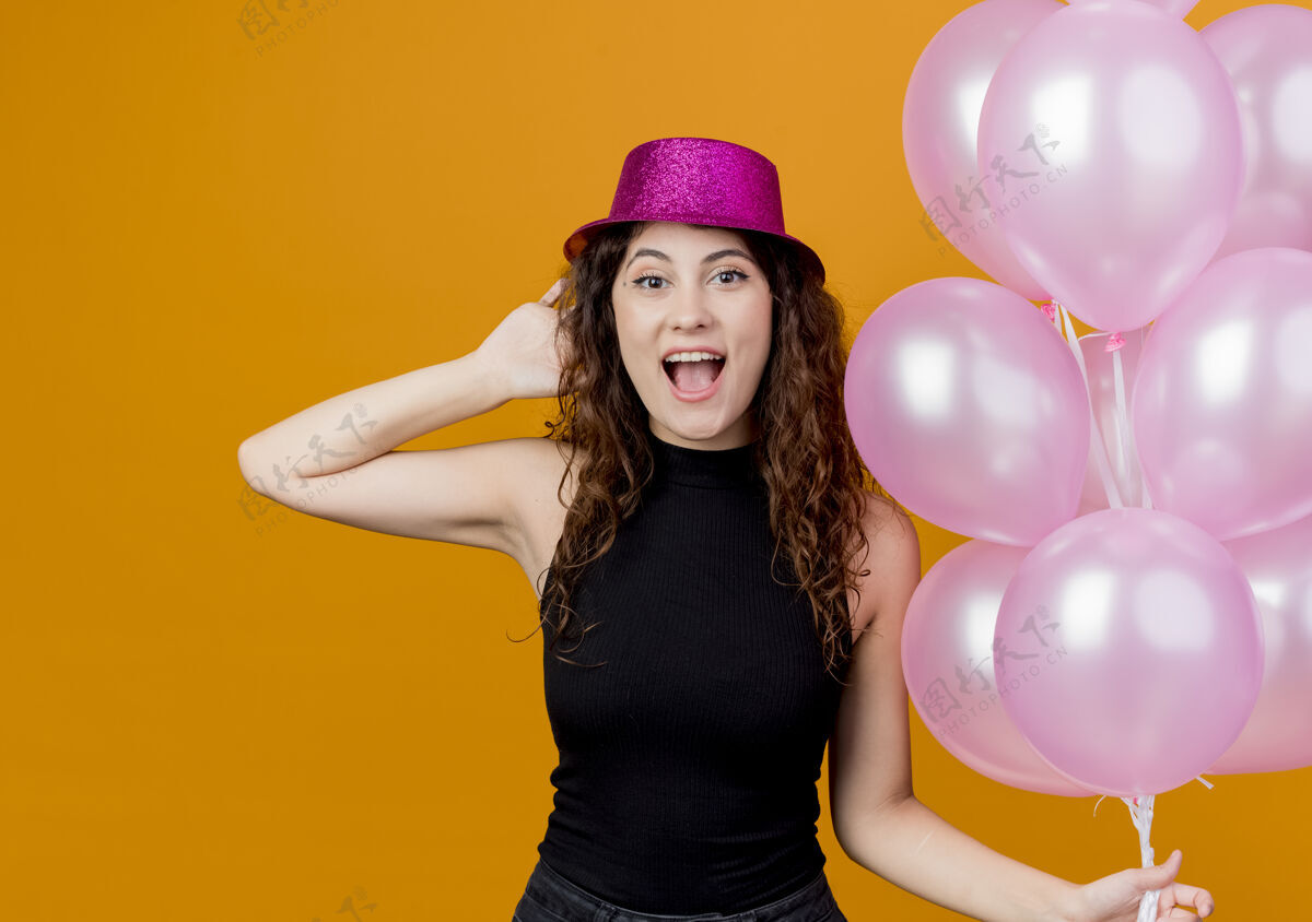 站着一位年轻漂亮的女士 卷发 戴着节日礼帽 手里拿着一束气球 站在橙色的墙上 快乐而兴奋地庆祝生日一群空气兴奋