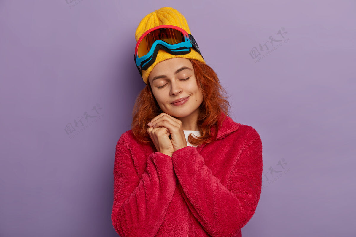 室内可爱的红发女人 喜欢冬天 歪着头闭上眼睛 穿着暖和的红色夹克 在紫色的墙上摆姿势困倦运动紫色