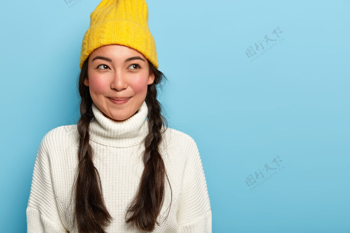 女人体贴迷人的亚洲年轻女子 笑容宜人 留着两条深色的辫子 身着冬装 蓝色背景 表达迷人的态度冬天帽子针织衫