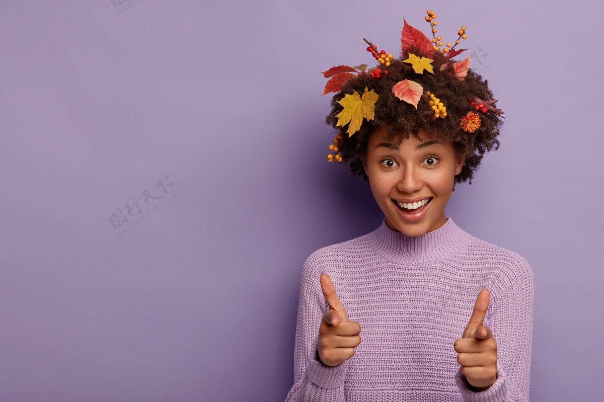 紫色室内喜色黑皮肤的女士镜头指着你 有着诱人的笑容 有着秋天的树叶扎在头发里 模特儿翻过紫色的墙壁手势毛衣手指