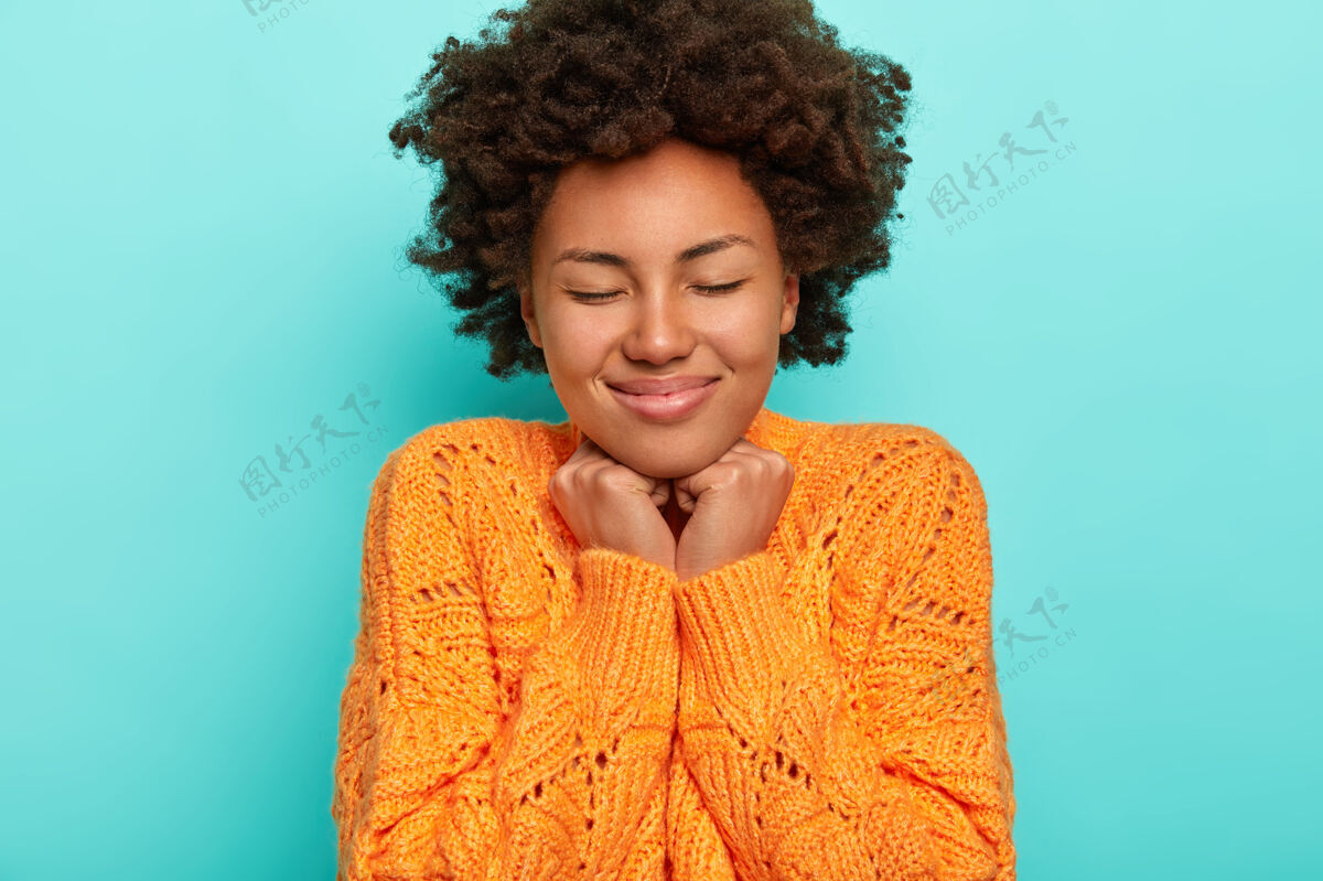 针织卷发女人的肖像 清爽的头发 手放在下巴下 感觉很愉悦 穿着针织橘色毛衣 隔离在蓝色背景上民族女人条纹