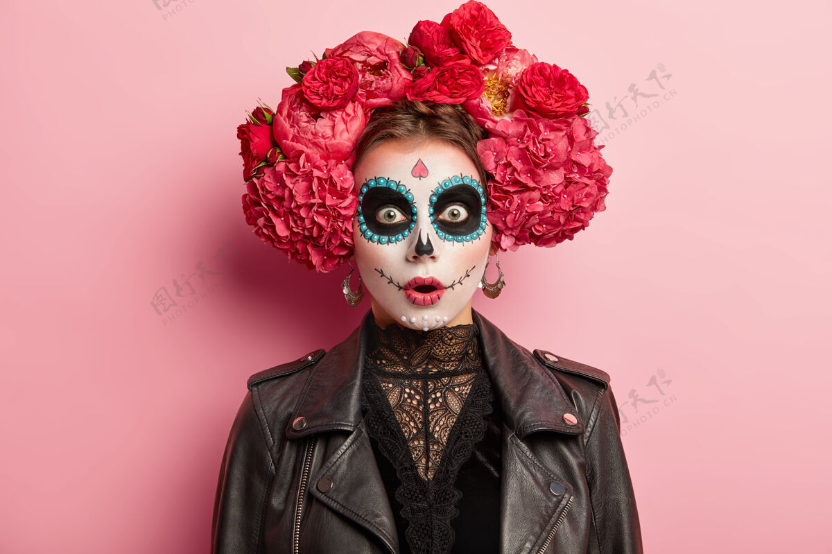 皮革惊魂未定的年轻女子有着吓人的鬼脸 为死亡节化妆 穿着黑色皮夹克 在玫瑰色的工作室背景上做模特骷髅女象征着死亡头像麻木身体