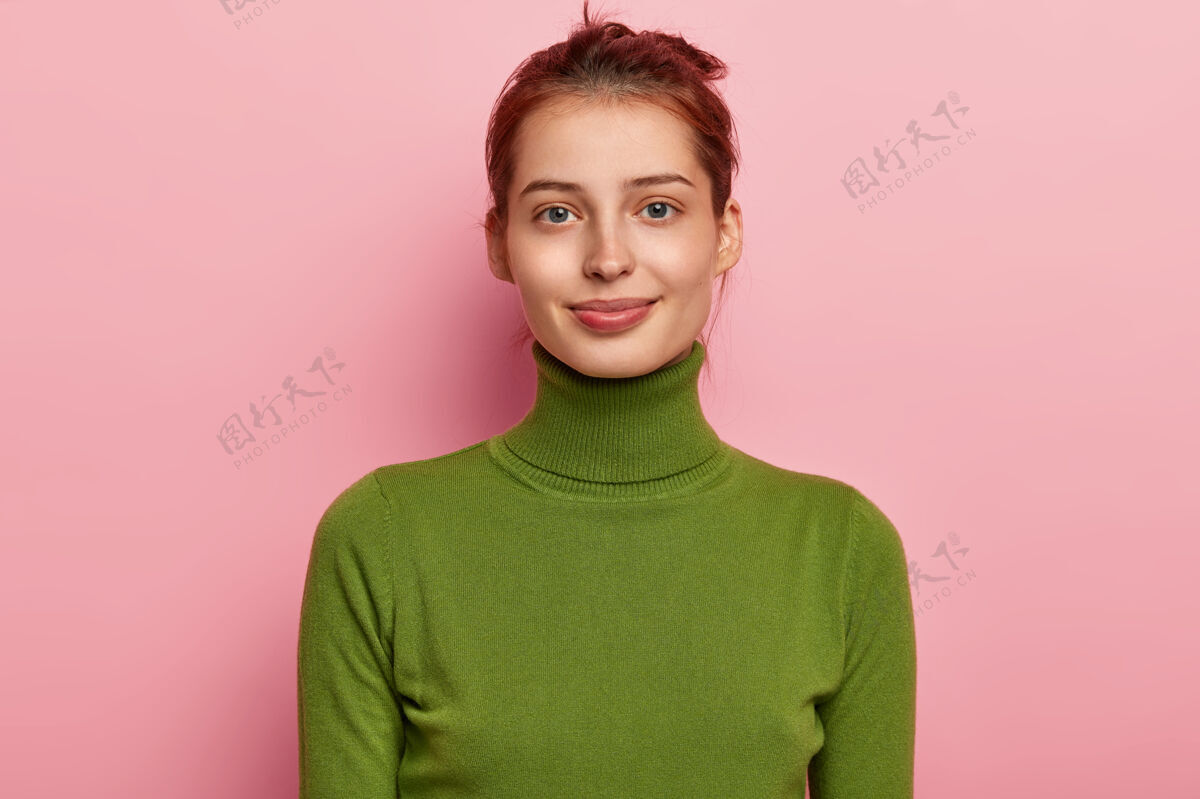 高兴美丽少女的肖像有深色的梳头 迷人的外表 穿着休闲的绿色高领毛衣 愉快地看着镜头 在粉色背景下摆姿势快递站年轻