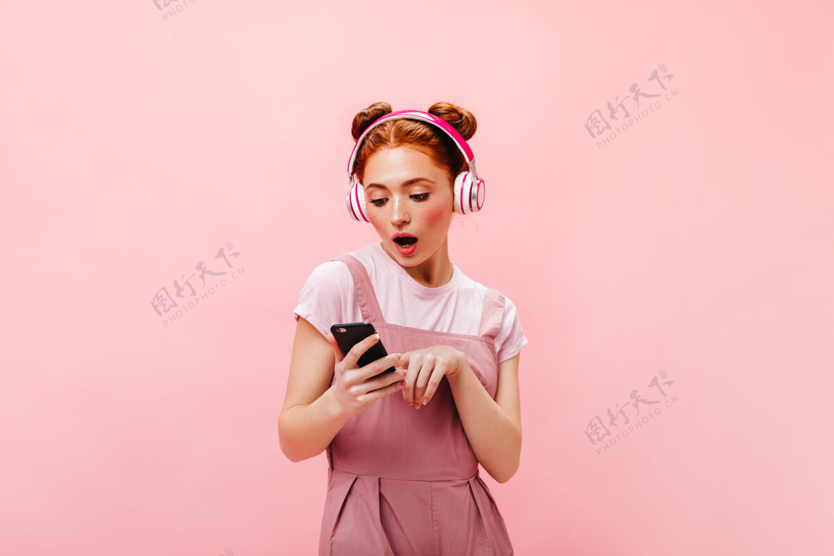 头带穿着粉红色裙子的惊讶的绿眼睛女人的肖像拿着智能手机和耳机听歌曲的女人优雅性感音乐