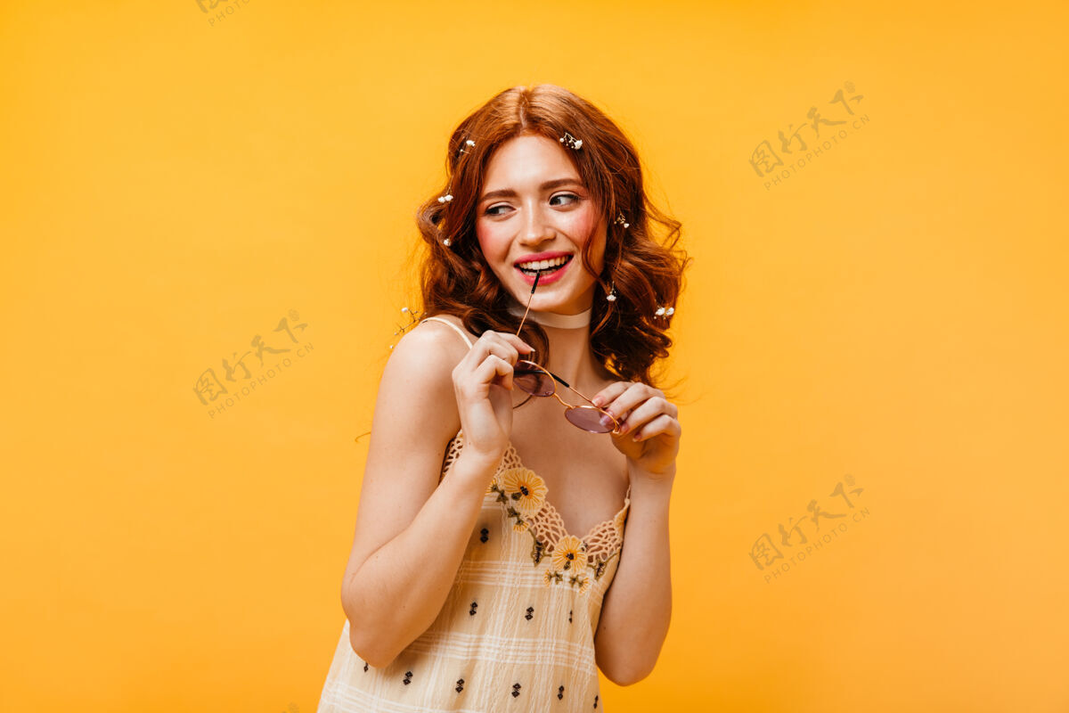 外观身着格子裙的红发女子微笑着咬着黄色背景上太阳镜的镣铐魅力太阳裙肖像