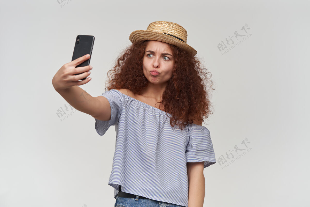 看起来十几岁的女孩 快乐的女人 卷曲的姜黄色头发穿着条纹露肩衬衫和帽子在智能手机上自拍 撅着嘴唇站在白色的墙上孤立模特手机自拍