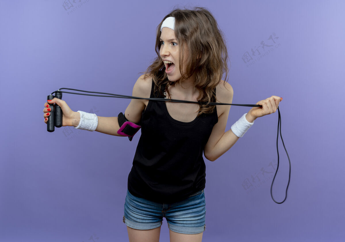 绳索身穿黑色运动服的年轻健身女孩 头戴钢带 手拿跳绳 站在蓝色的墙上 吓得站在一边看靠边女孩看