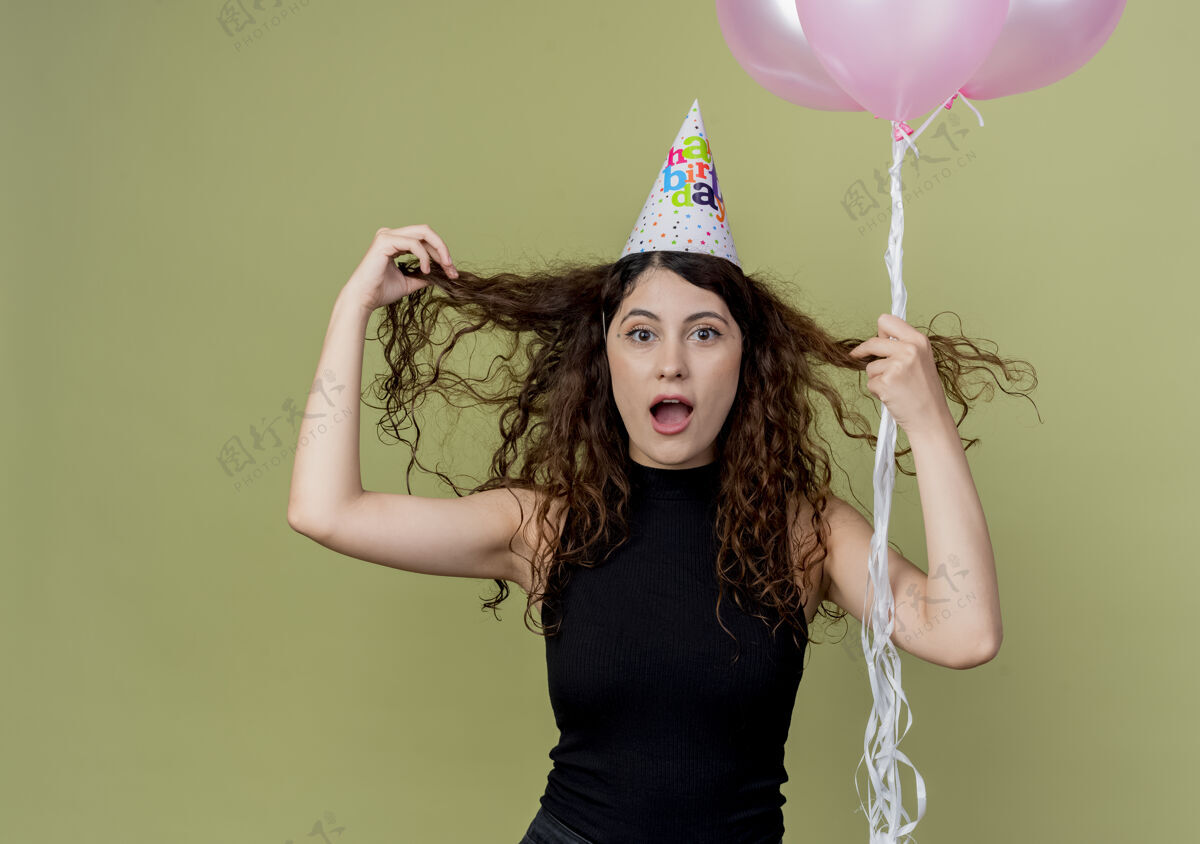 站着一个年轻漂亮的女人 一头卷发 戴着节日帽 手里拿着气球 困惑和惊讶地站在光墙上的生日派对概念卷曲气球抱着