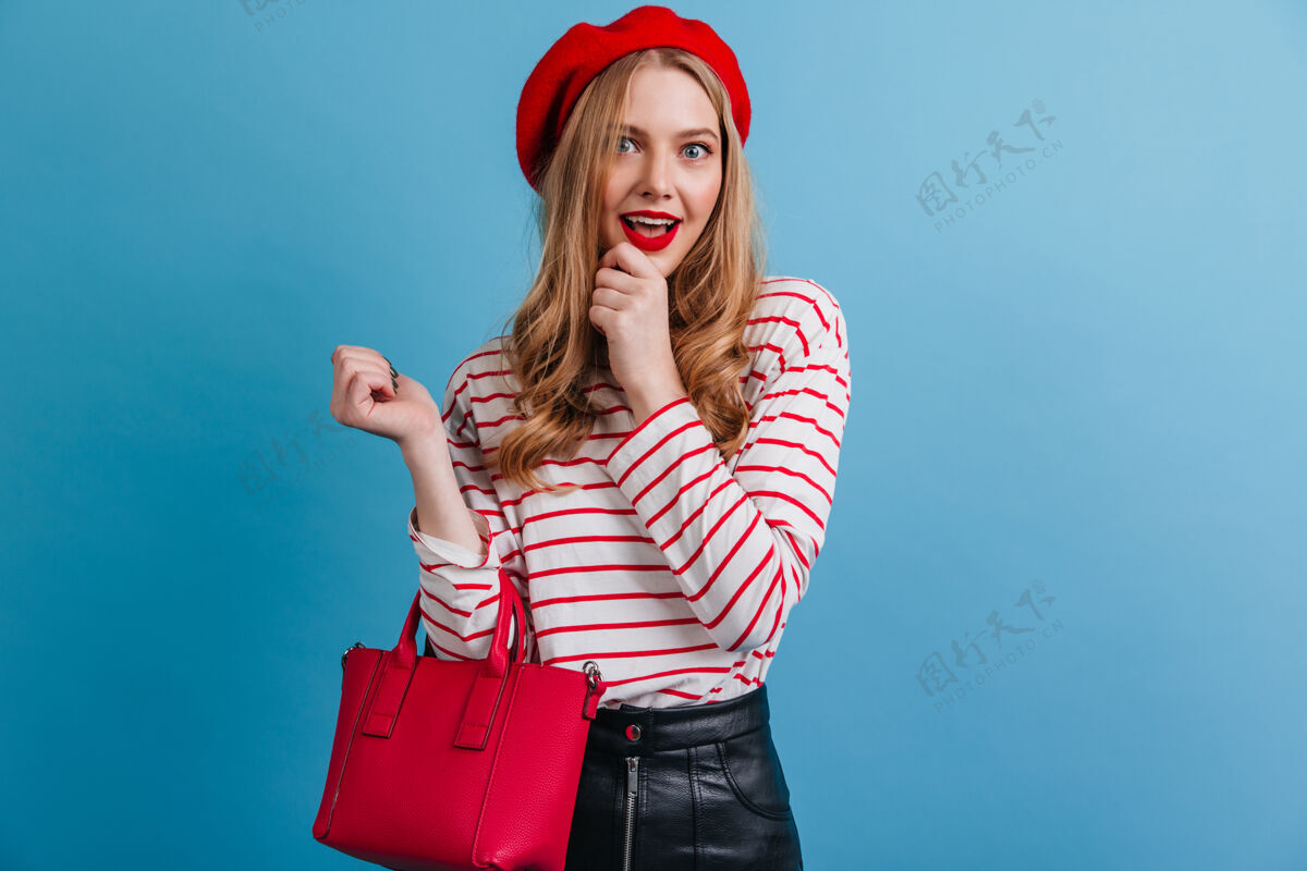 时尚穿着红色贝雷帽的迷人女孩蓝色墙壁上孤立的法国金发女郎的正面视图情绪帽子欢乐