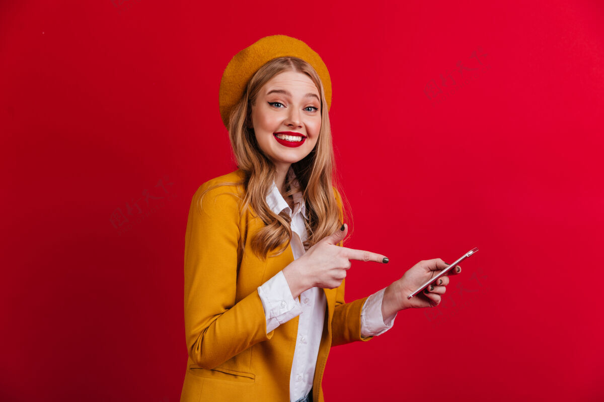 数码设备开朗的法国女士用手指着智能手机微笑着戴贝雷帽的金发女孩站在红墙上年轻时尚年轻人