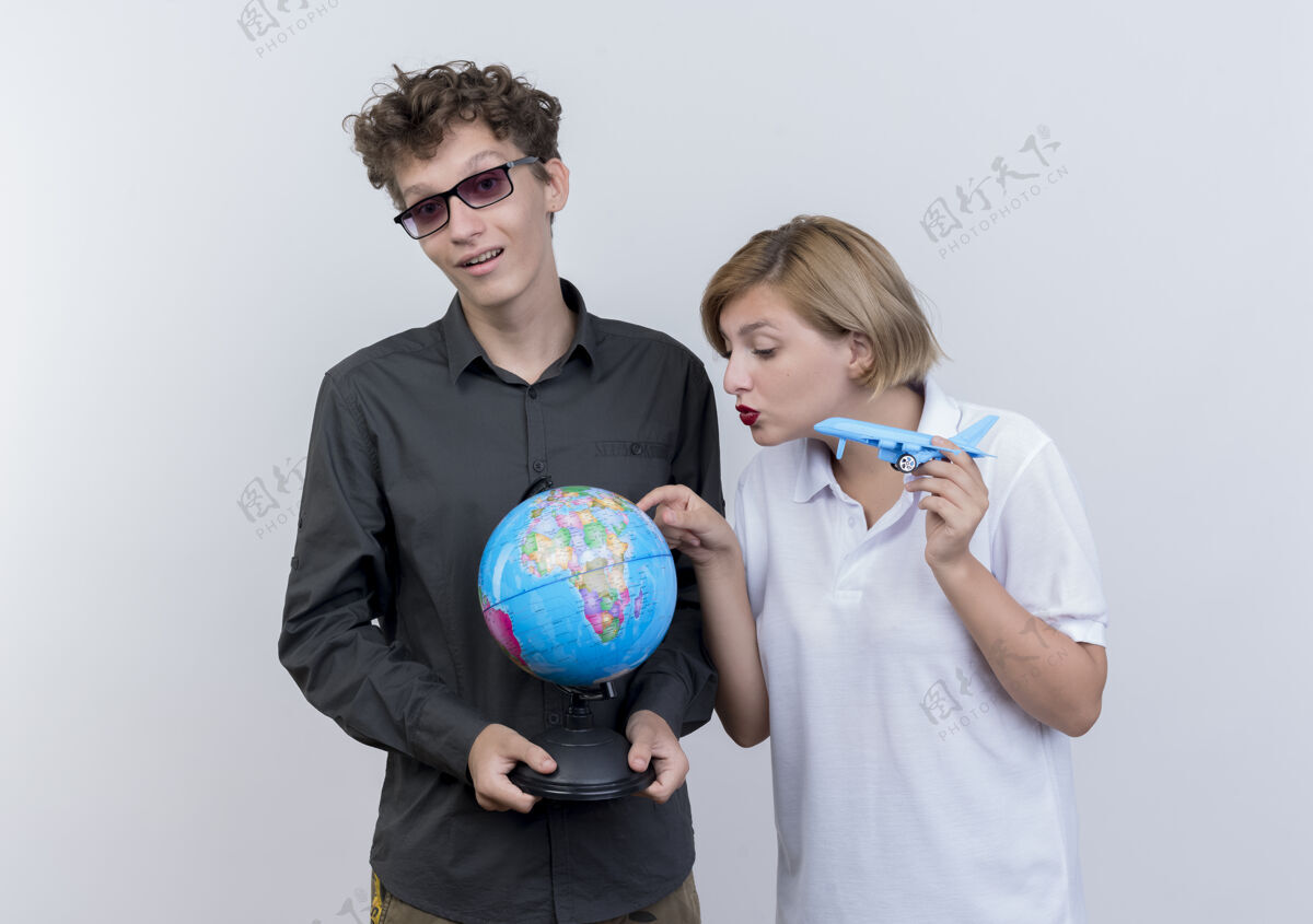 抱着一对幸福的年轻游客男女手持地球仪和玩具飞机一起站在白墙上情侣站着男人