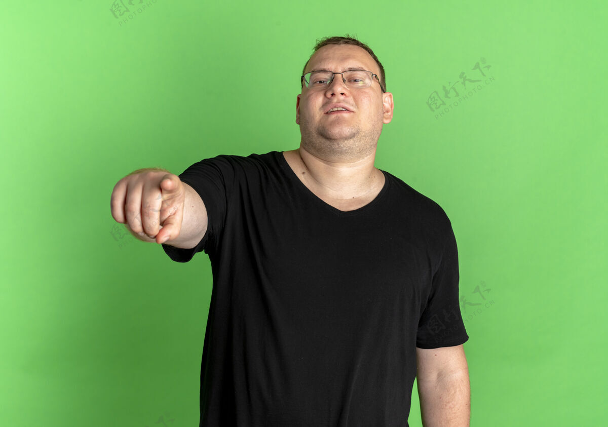 眼镜戴着眼镜 身穿黑色t恤 手指微笑 站在绿色墙壁上的一个高兴的超重男子站着请戴超重