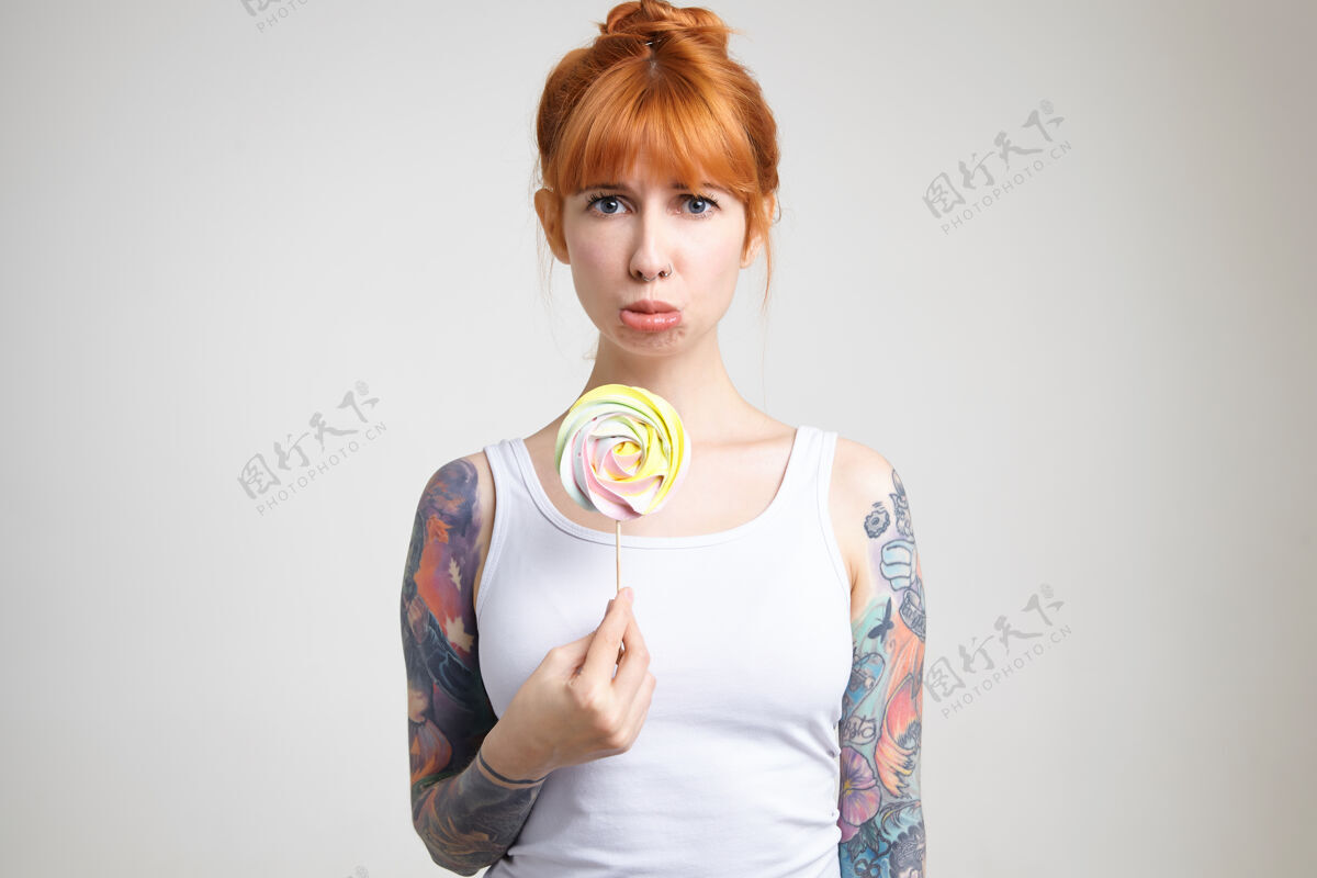 姿势得罪了年轻漂亮的红发女士 她身上有纹身 手里拿着糖果 穿着白衬衫站在白色背景上 悲伤地看着镜头衬衫情绪休闲