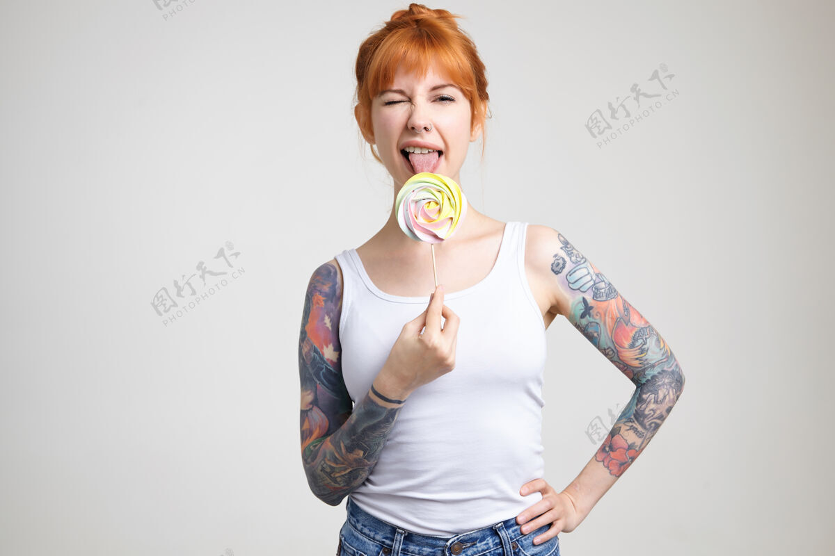 红发摄影棚拍摄的一张照片 年轻快乐的纹身女性 狐狸精的头发伸出舌头 一边舔糖果 一边对着镜头眨眼 隔离在白色背景下成人白皙结