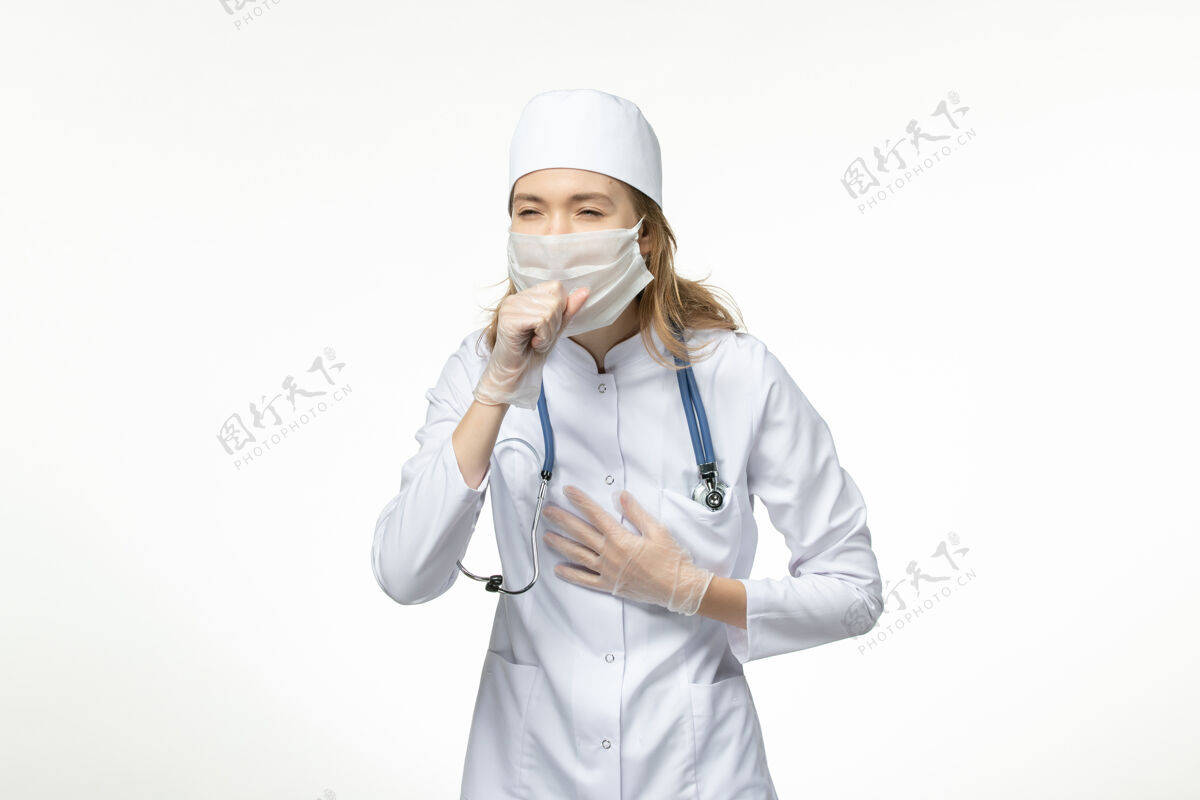 大流行正面图年轻女医生因冠状病毒在白色表面咳嗽 身穿医疗服 戴防护口罩正面到期听诊器