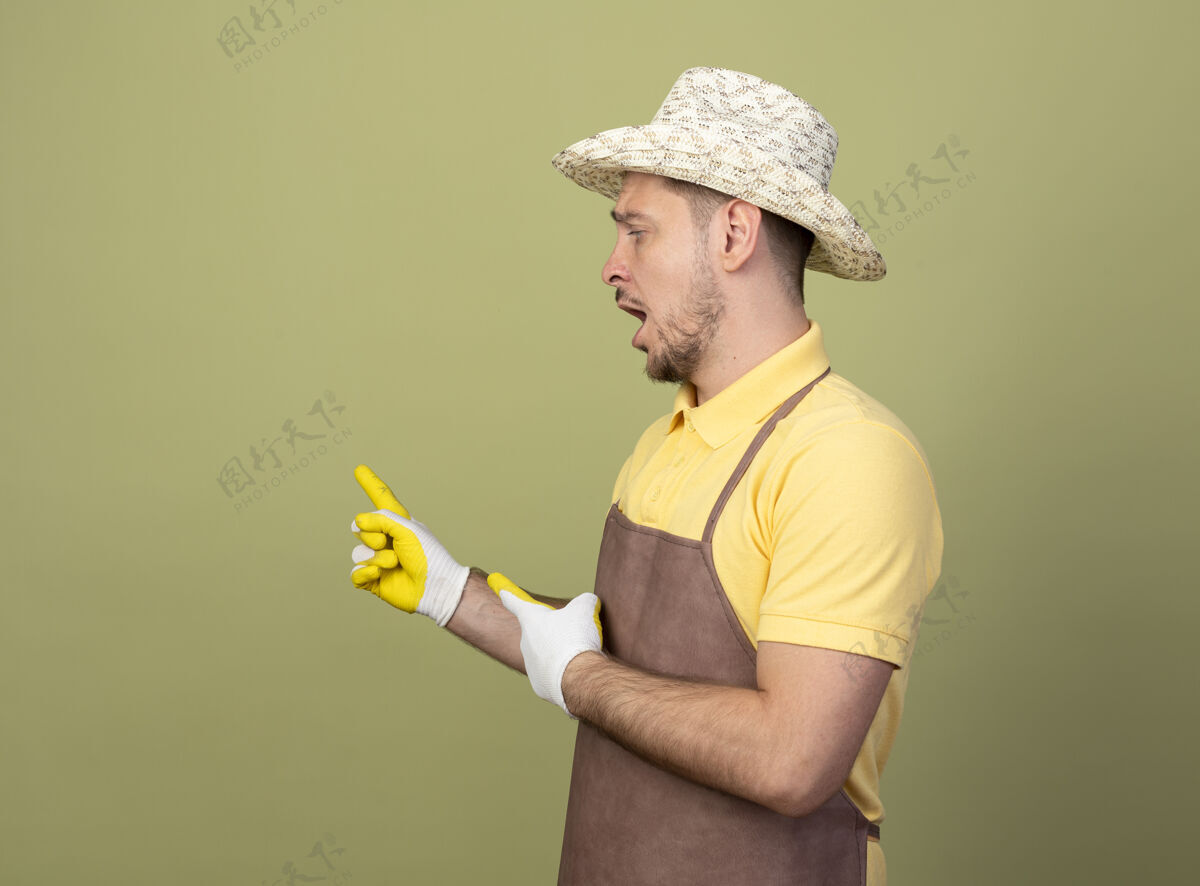 手套年轻的园丁穿着连体衣戴着帽子戴着工作手套用食指指着背看着困惑花园帽子站