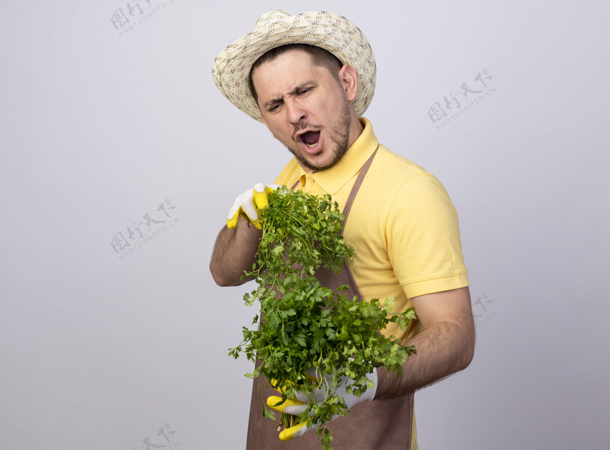 帽子年轻的园丁穿着连体衣 戴着帽子 戴着工作手套 手里拿着新鲜的草药 高兴而兴奋草药手套年轻